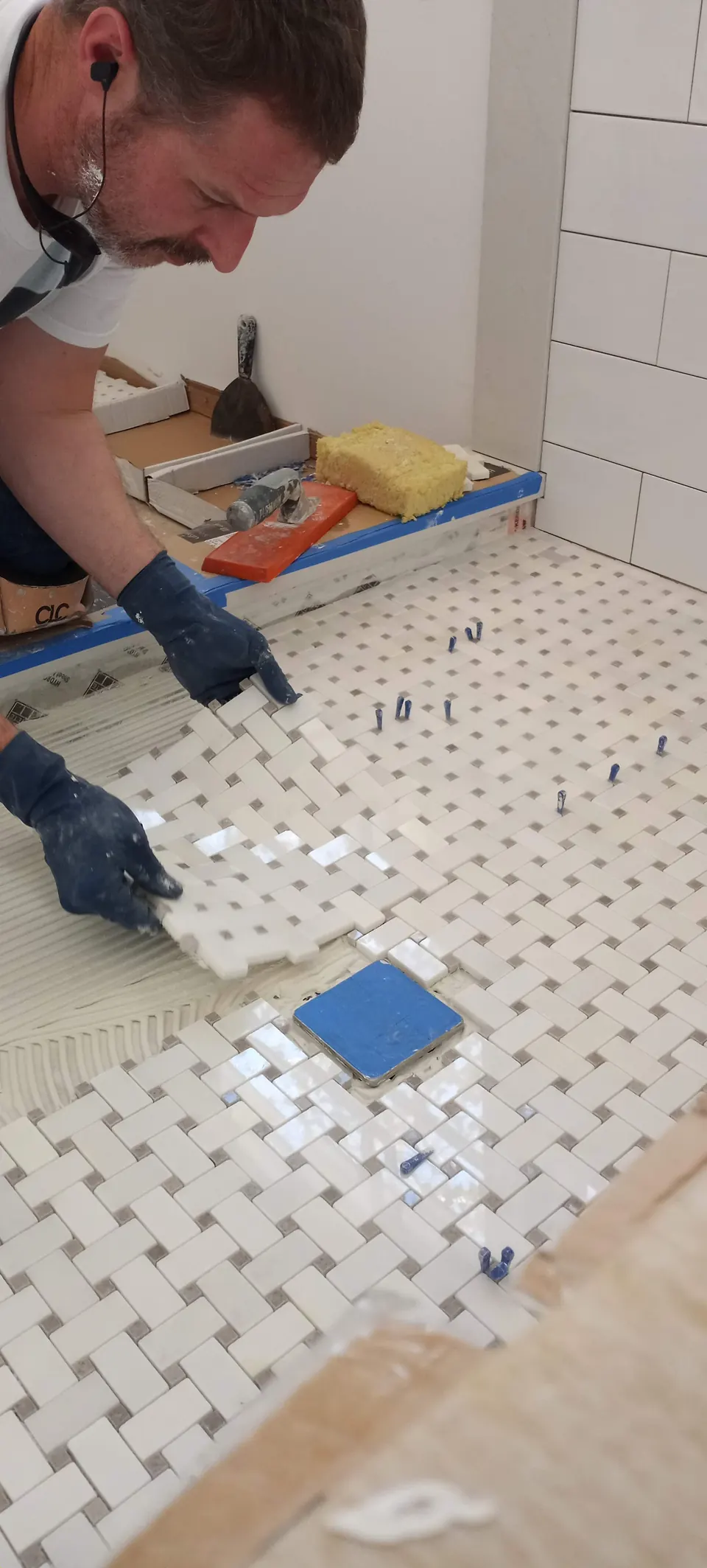 Brian setting white marble basketweave mosaic on a shower floor, with leveling clips and the drain covered