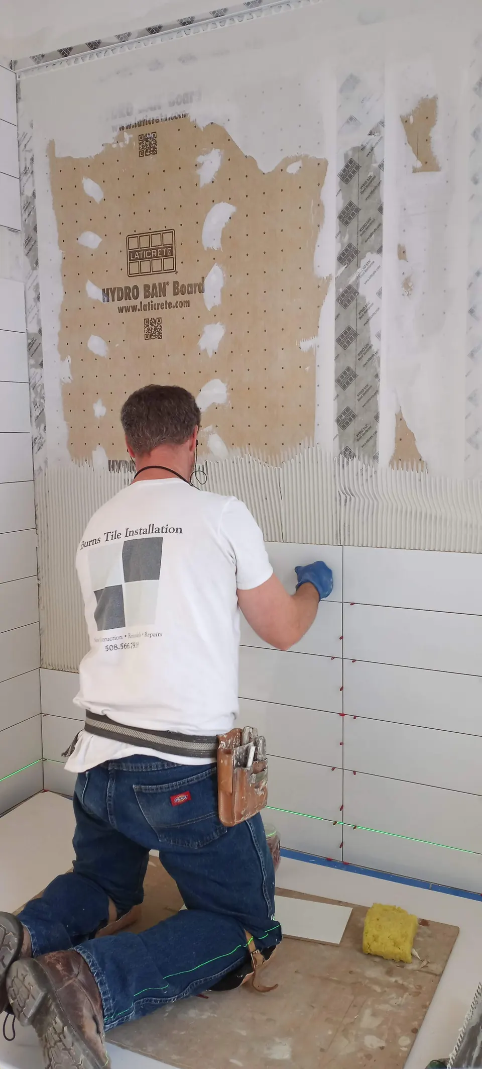 Brian kneeling in a shower to set large white wall tile on waterproof foam board, following laser level lines