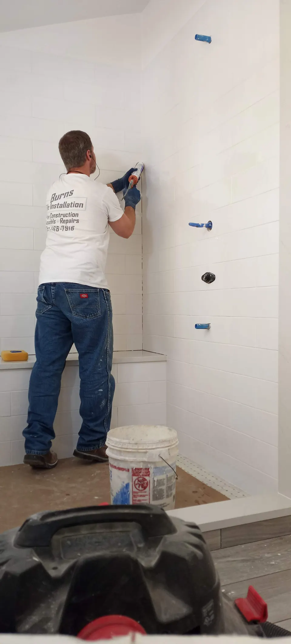 Brian working on a large white tile shower wall with a bench, a bucket and a shop vacuum in the foreground