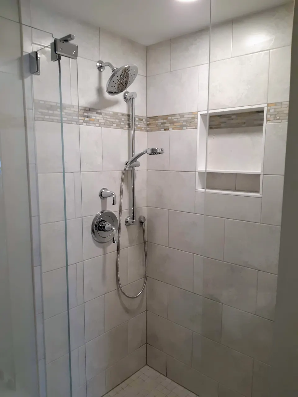 Walk-in shower with large light gray wall tile, a glass and stone mosaic accent band, a recessed niche and a frameless glass door