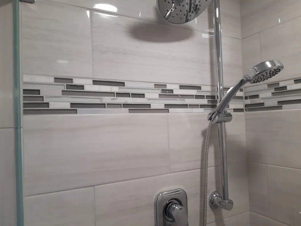 Close-up of a glass and stone linear mosaic band set between large shower wall tiles, beside a chrome hand shower