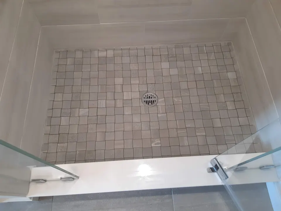 Shower floor of tumbled stone square mosaic sloped to a center drain, with a white curb and glass door hardware