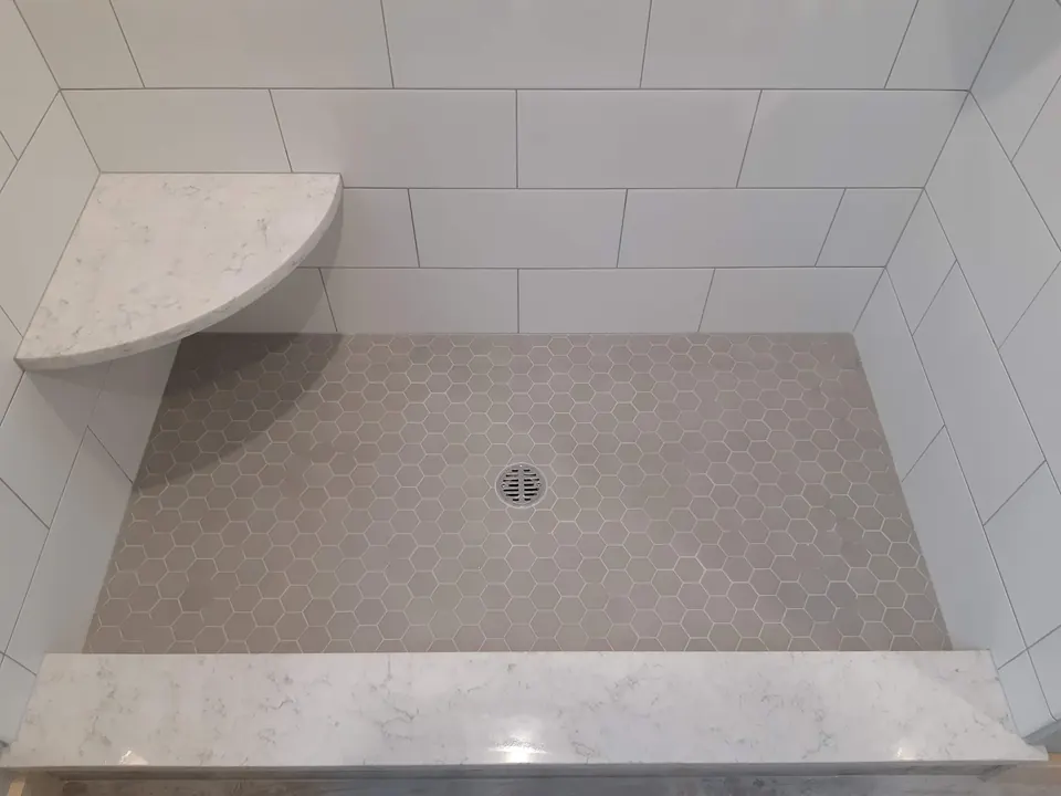 Gray hexagon mosaic shower floor with a center drain, a stone corner seat and a stone curb, surrounded by white wall tile