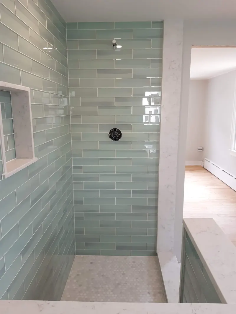 Finished walk-in shower in seafoam glass subway tile with a recessed niche, a half wall and a white hexagon floor