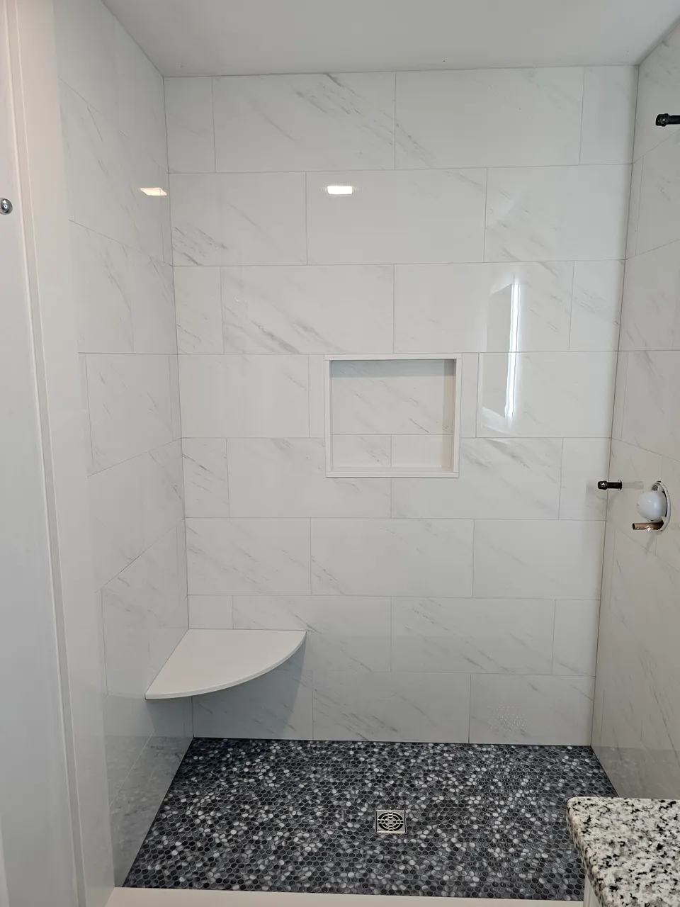 Walk-in shower with large white marble-look tile, a recessed niche, a corner seat and a black and white pebble mosaic floor
