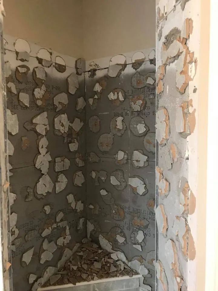 Shower stall stripped of tile, showing spot-bonded adhesive dabs on the backer board and broken tile on the floor